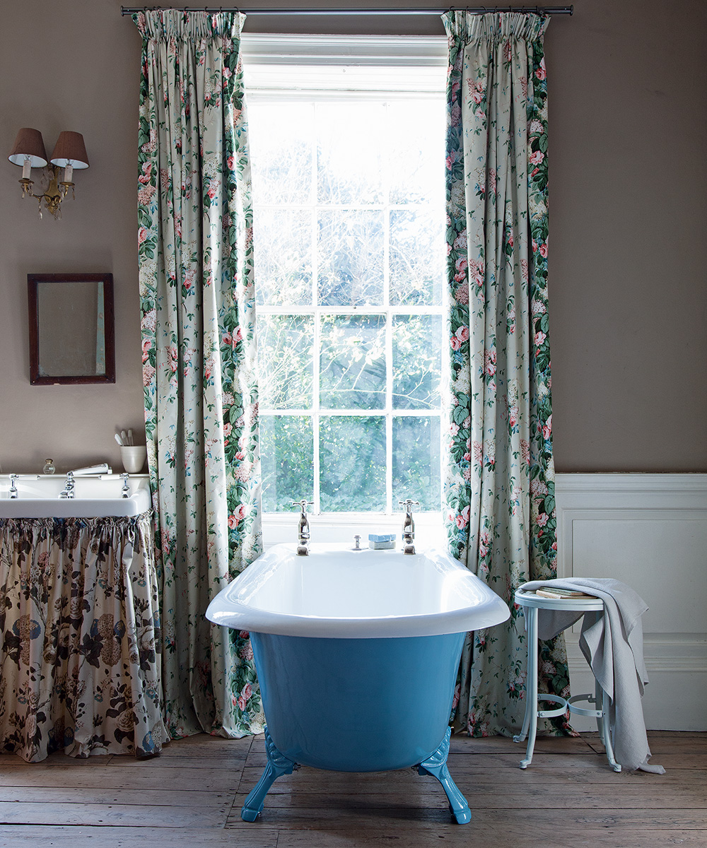 A blue free-standing bath in front of a window with floral curtains