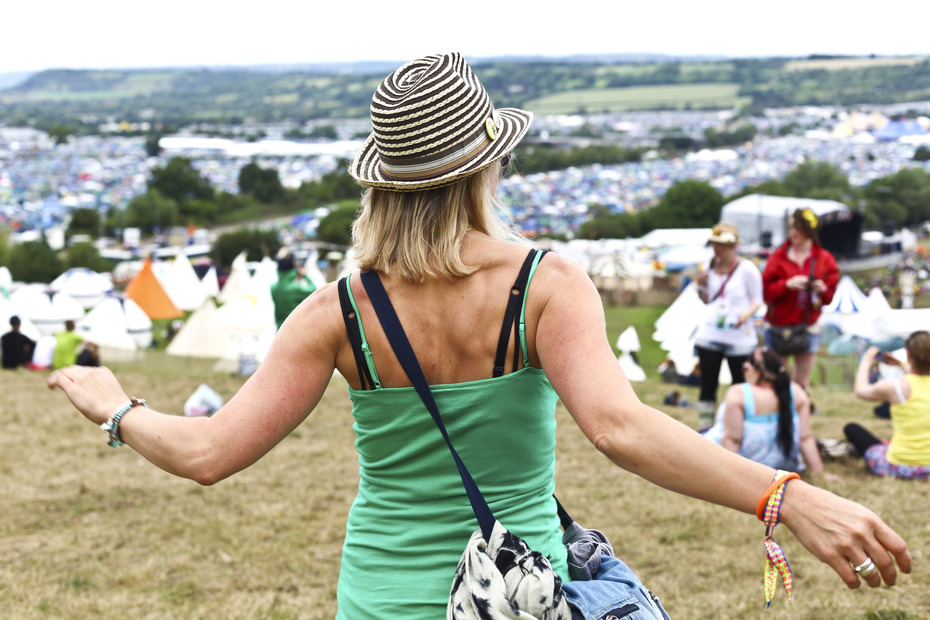 A woman in a green vest and straw hat is standing over the Glastonbury fields filled with tents.