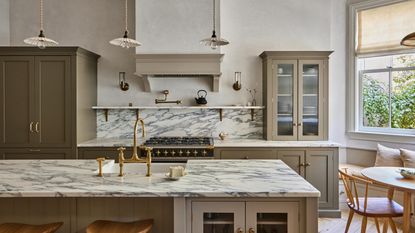 A khaki style kitchen and on the right side of the kitchen is a fluted glass cabinet. In the kitchen is also a large marble island with a marbled backsplash.
