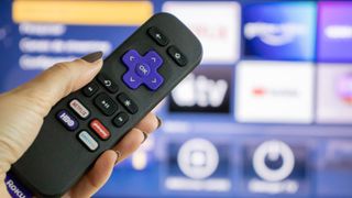 A hand holds a Roku remote in front of a TV with the Roku home screen.