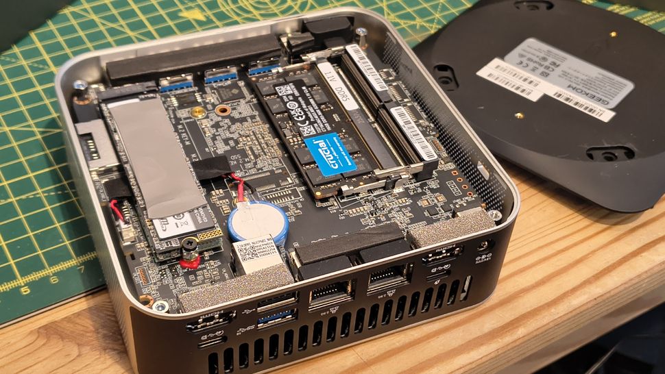 Open computer case showing internal components like RAM, SSD, and circuit board. The cover is off to the side, revealing ports and hardware details, on a wooden surface.
