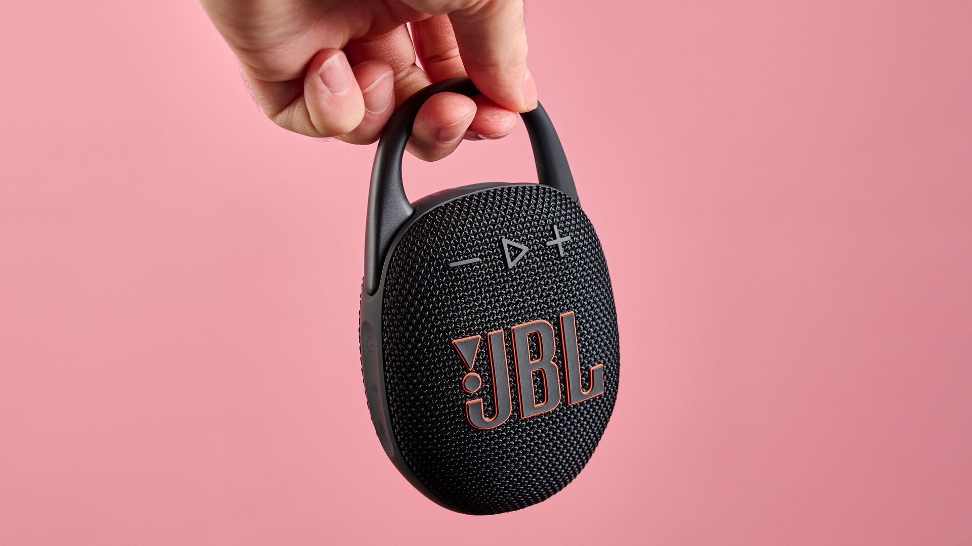 The JBL Clip 5 in black is pictured being held by the carabiner against a pink background,