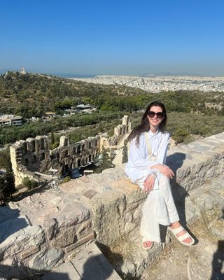 Anne Hathaway sitting on a cliffside wearing a white linen button-down shirt with linen pants and white Birkenstock Arizona sandals.