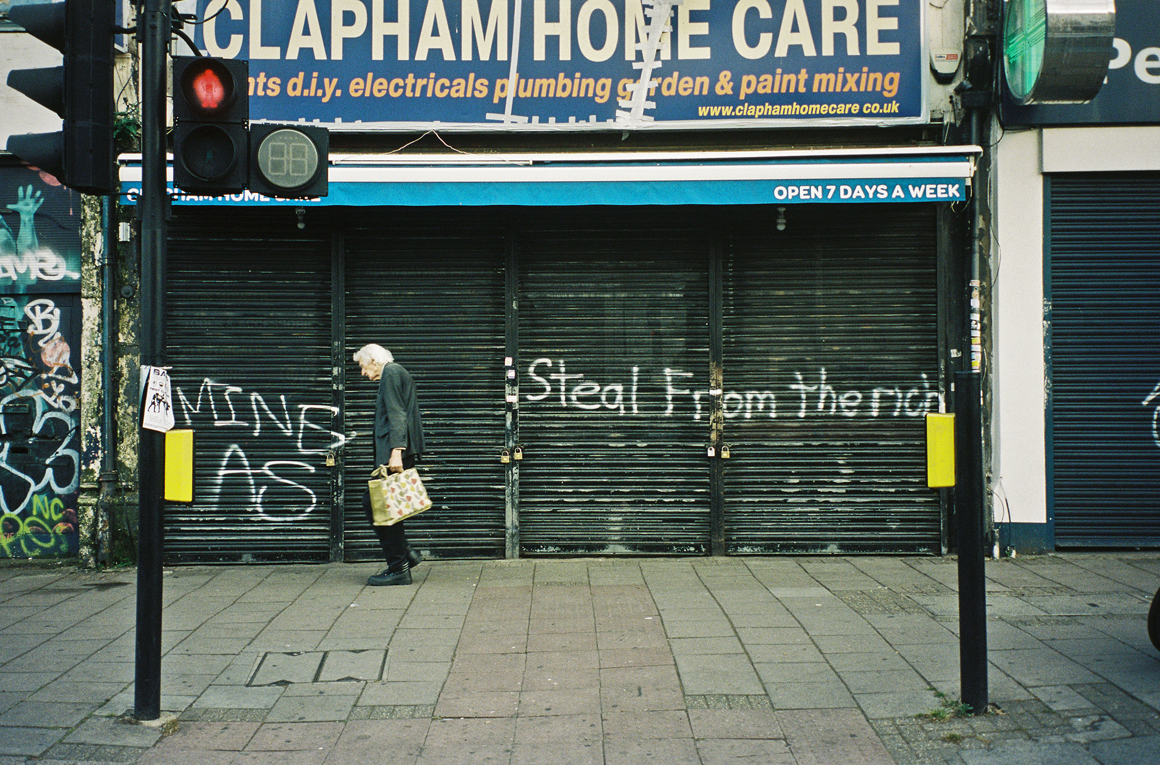 Colour photograph of an elderly person walking along the street past a shuttered shop front that has “STEAL FROM THE RICH” spray-painted in front