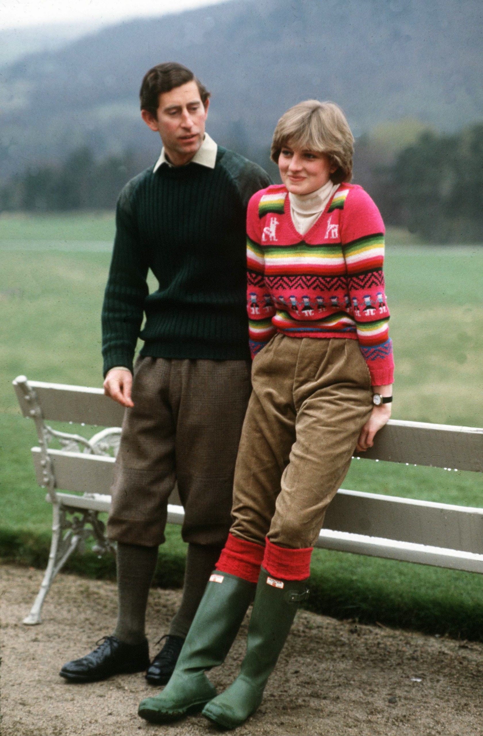 Princess Diana leaning against a bench wearing a bright sweater and rain boots with Prince Charles