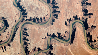 An astronaut photo of a winding grenn-color river covered partly by shadows from canyon walls