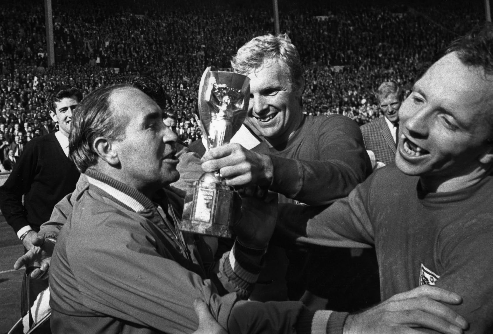 England Manager Alf Ramsey (left) celebrates his team's 4-2 victory in extra time over West Germany in the World Cup Final at Wembley Stadium. With him is captain Bobby Moore (1941 - 1993), holding the Jules Rimet Trophy, and team mate Nobby Stiles, 30th July 1966. (Photo by Hulton Archive/Getty Images)