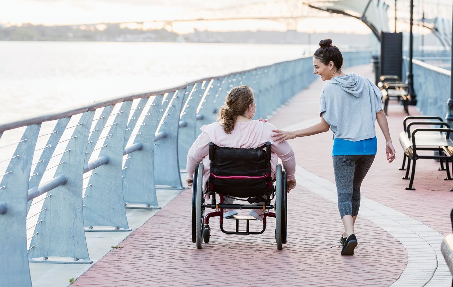 A woman in a wheelchair is being assisted by another woman along a scenic walkway, highlighting themes of support and accessibility.