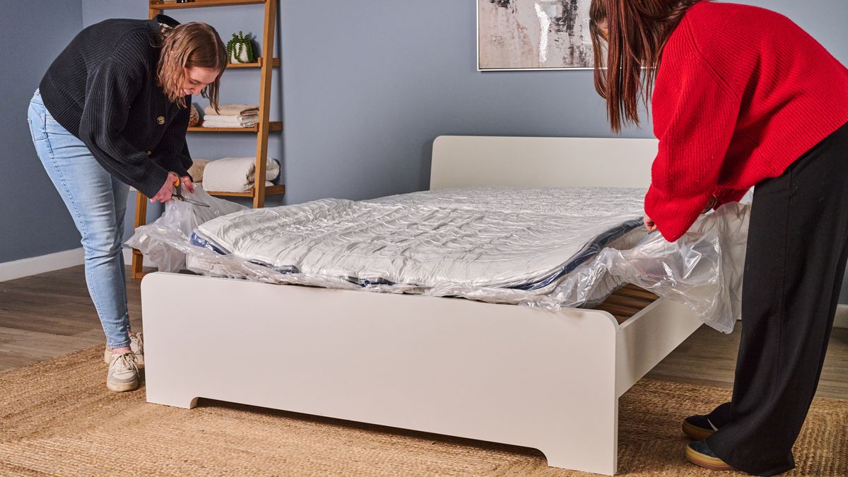 The image shows two women taking the Helix Midnight mattress out of its packaging and onto a white bed frame in a blue bedroom