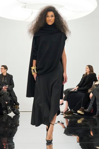 Photo from the F/W 24 Alaïa show of a brown-haired model wearing a high-neck asymmetric black dress with a gold cuff bracelet and black round-toe heels.