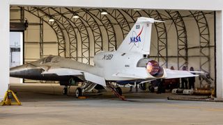 a white jet sits in a hangar with a cone of orange fire blasting from a cone-shaped nozzle on its tail