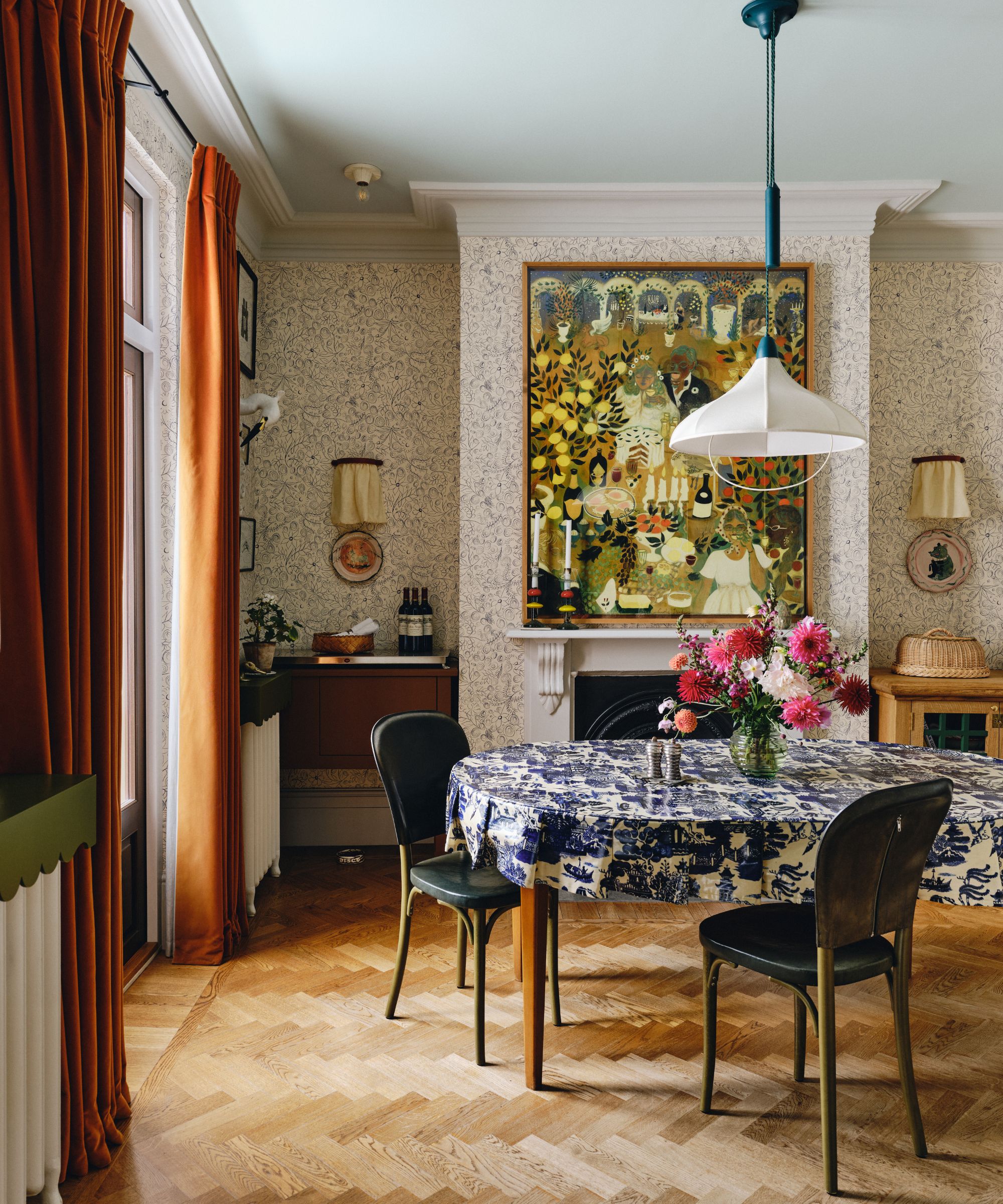 scandi maximalist style dining room with wallpapered walls, a large fireplace with folk art hung above, dark orange velvet curtains, a dining table with blue and white printed tablecloth and a white fabric pendant light