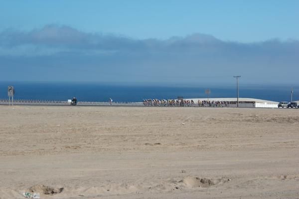 The peloton rolls along the desert next to the sea.
