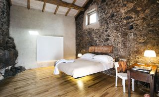 A hotel bedroom with a bed, a desk, wooden floors and rough dark stone walls.