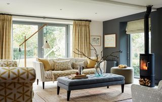 country-style living room extension to converted barn with wood-burning stove and grey walls, with beige sofas and beige curtains and doors leading out to garden