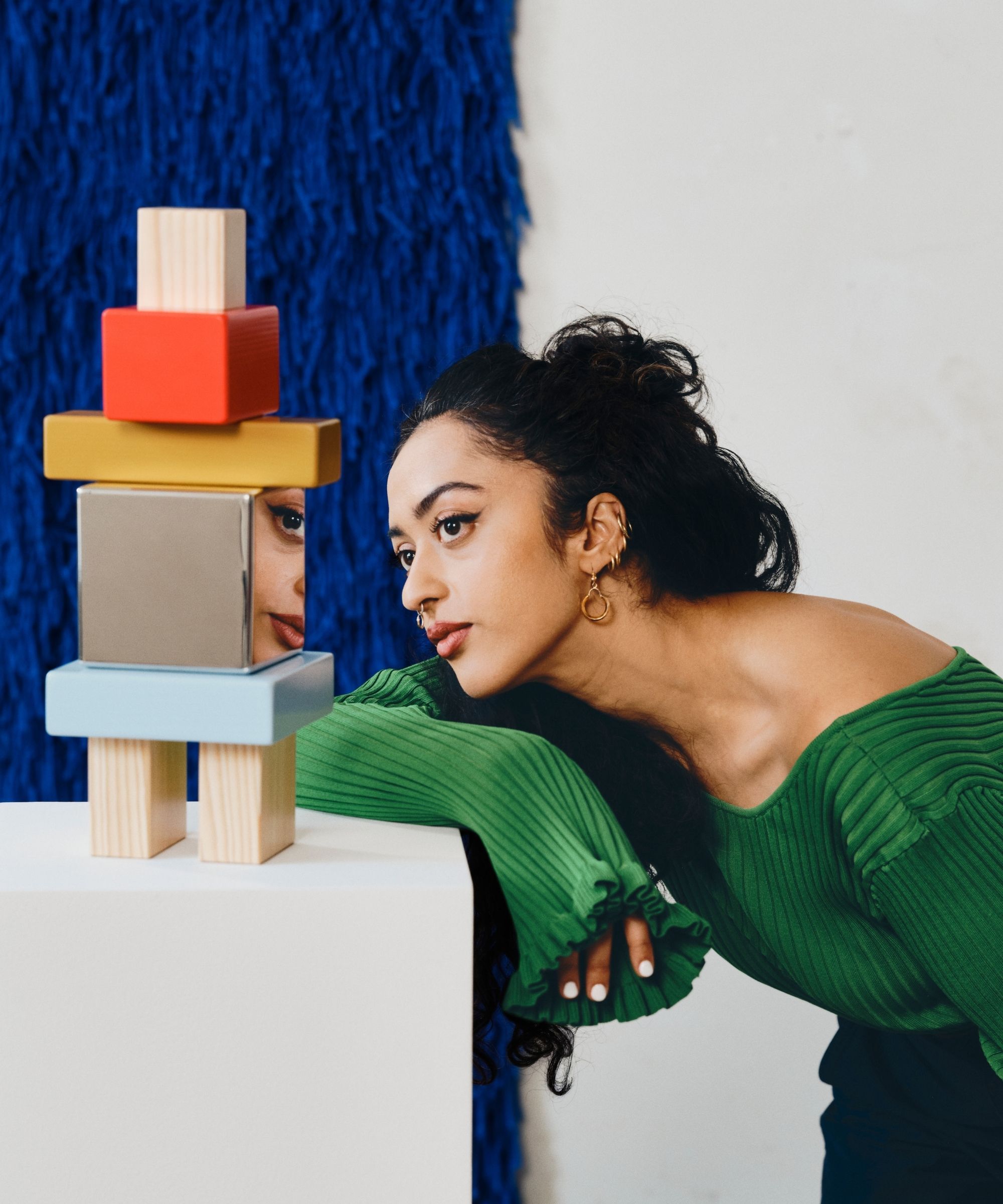 girl in green top looking at a colorful stack of blocks on a white plinth in front of a shaggy blue wall hanging