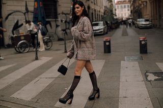 Woman at Paris Fashion Week wearing sheer knee-high socks, black platform heels, and a plaid blazer walking in the street.