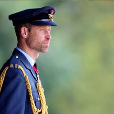 Prince William standing against a green background wearing a military uniform and smirking