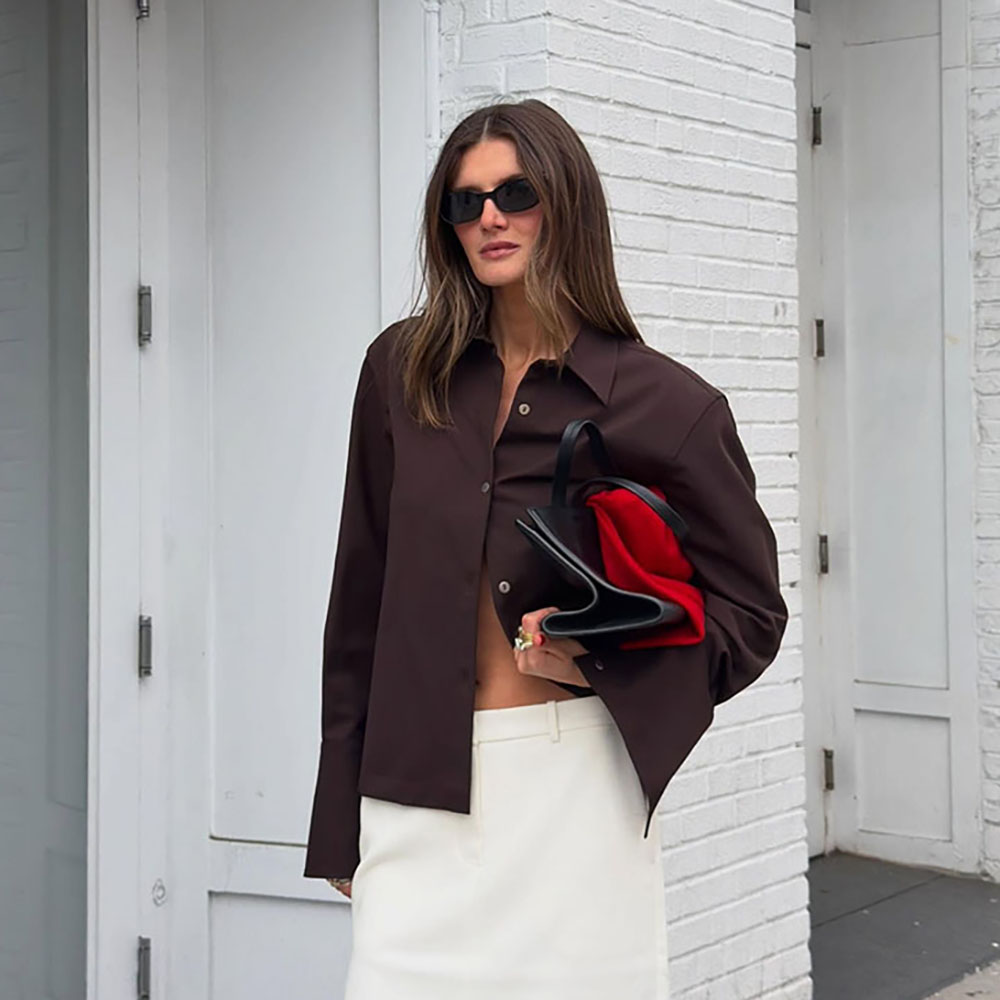 a woman wearing a brown button-down, white skirt, black clutch, and black sandals