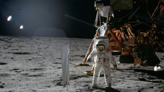 An astronaut wearing a white space suit stands next to a flag on the moon with a large golden lunar lander behind him