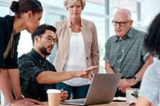 Office workers of different ages and generations gather around a computer for an informal meeting.