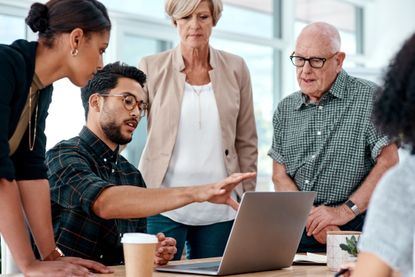Office workers of different ages and generations gather around a computer for an informal meeting.