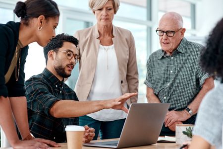 Office workers of different ages and generations gather around a computer for an informal meeting.