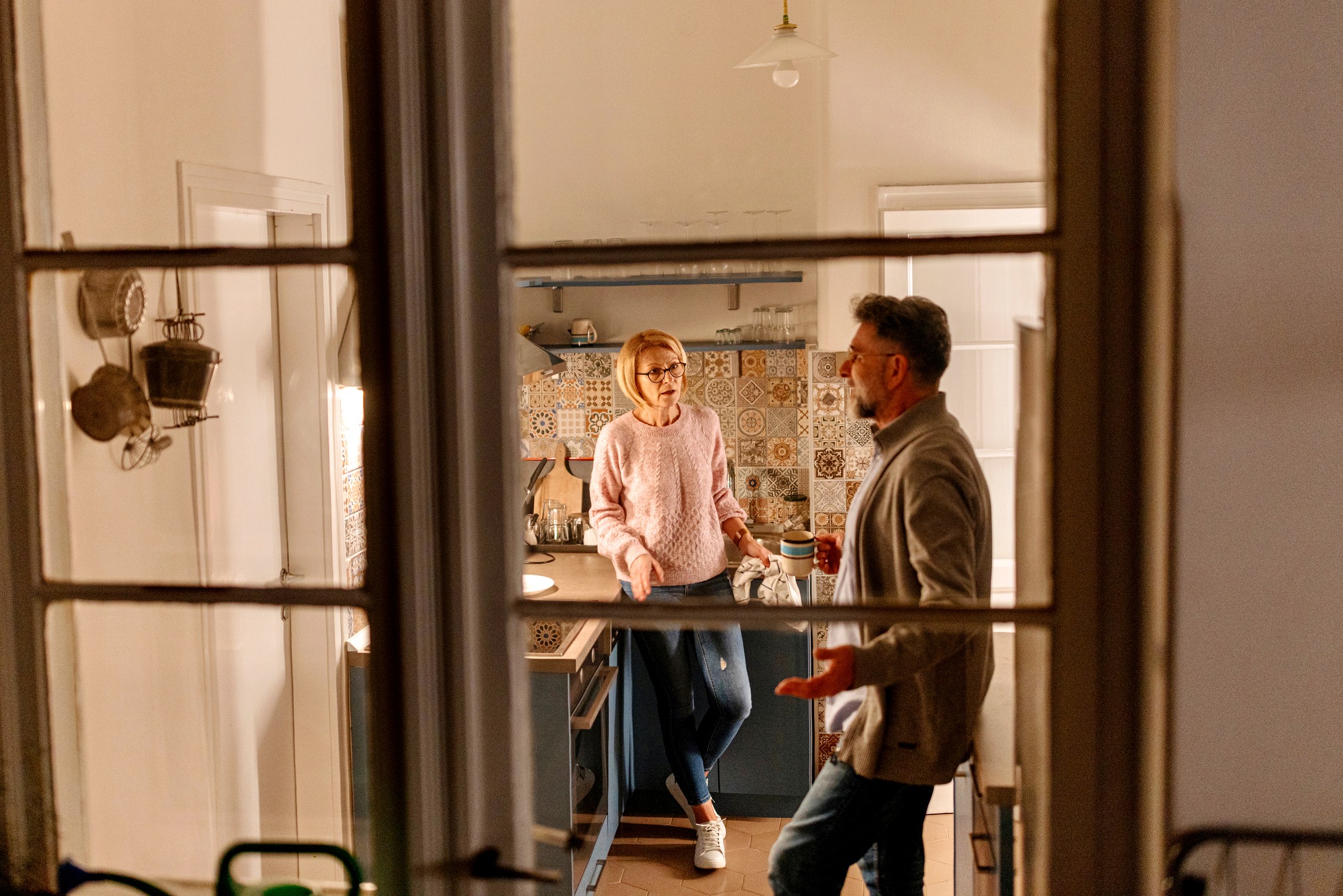 View of a senior couple in their kitchen having an argument. The viewer is looking through the window from outside the house.