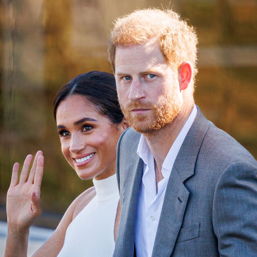 Meghan Markle wears a white halter top and waves while Prince Harry, who has red hair, wears a gray suit jacket and white shirt
