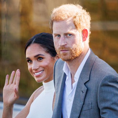 Meghan Markle wears a white halter top and waves while Prince Harry, who has red hair, wears a gray suit jacket and white shirt