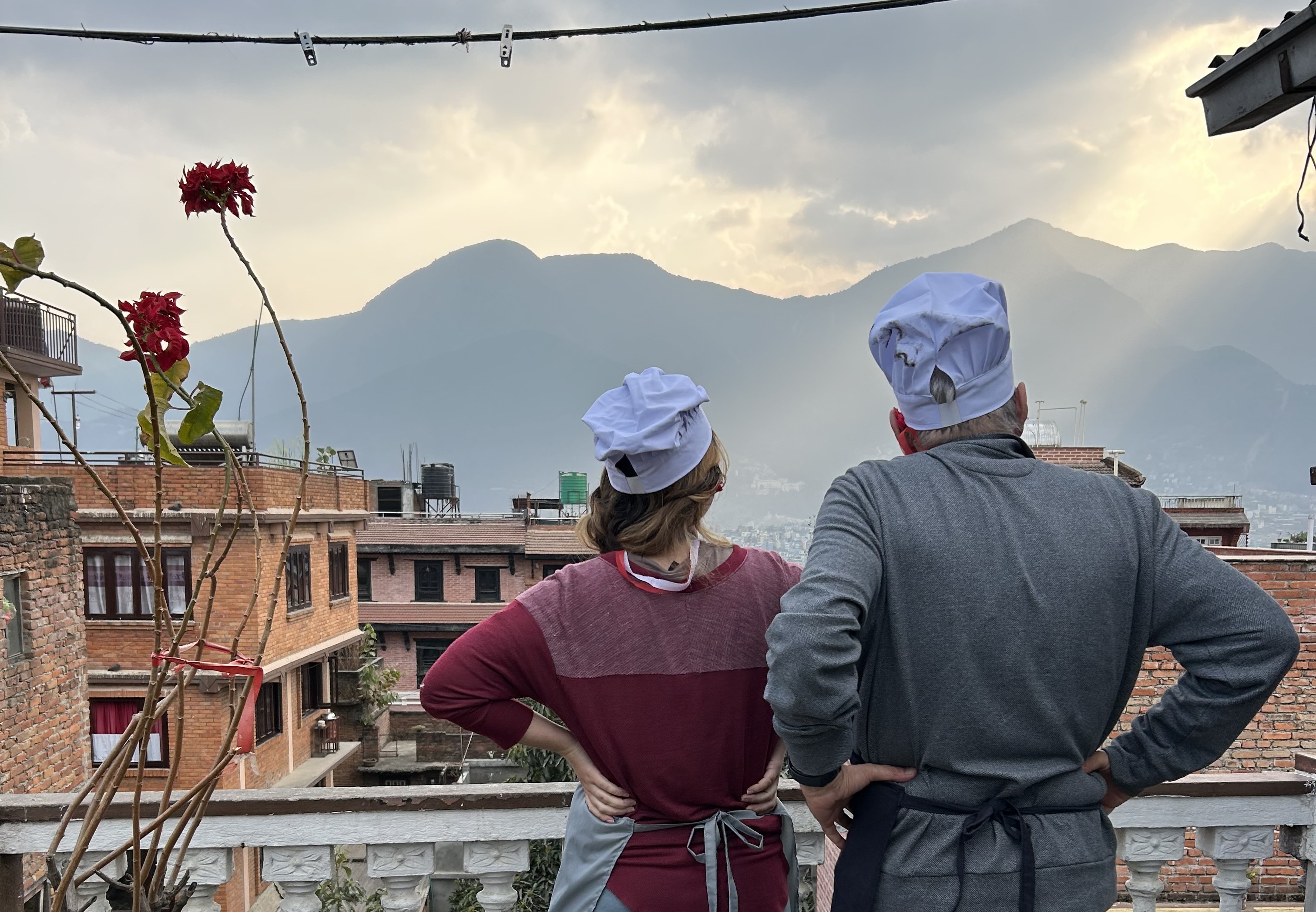 A woman and her father make the same pose with hands on hips looking out over a balcony in Kathmandu, Nepal, wearing chef's hats.