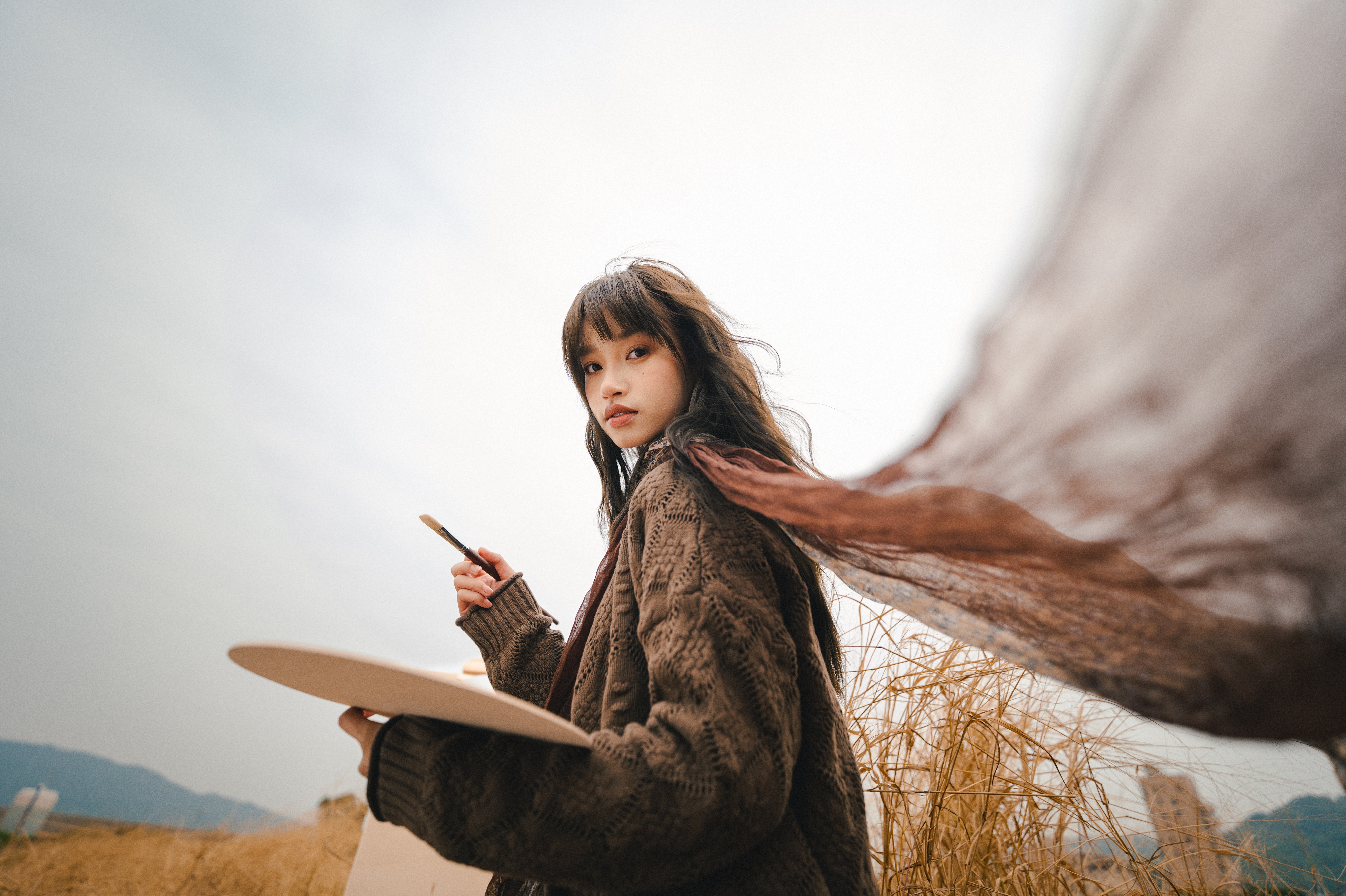 Woman painting from an easel in a field as her scarf billows in the wind