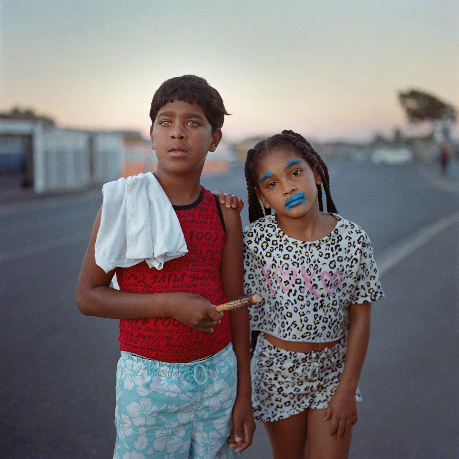 Two young children stand side by side on an asphalt road at dusk, with one wearing blue face makeup.