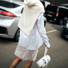 A woman wears a cream knit knotted around her shoulders, a white shirt, white bag and white skirt