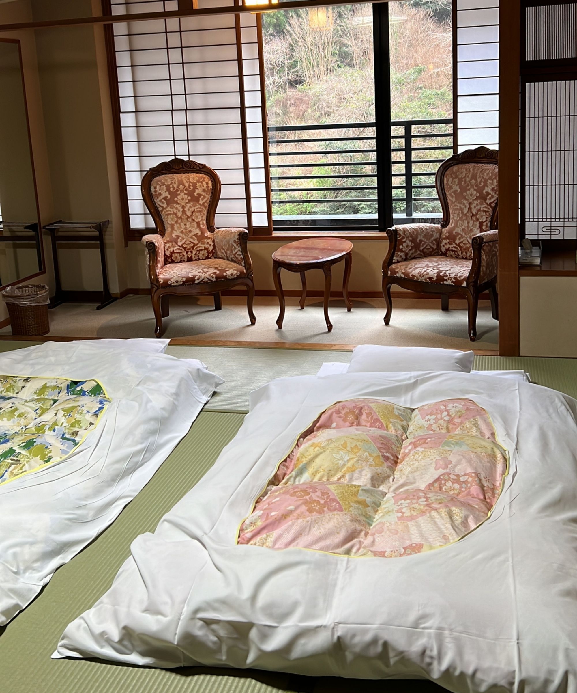 A traditional Japanese ryokan bedroom with two futon beds laid out on tatami mats, with a seating area and large windows seen behind