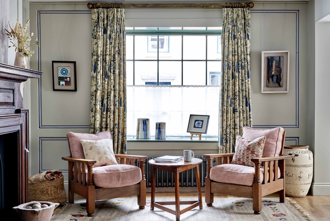 A living room with two arm chairs, a sage green wall with a wallpaper border, and patterned curtains