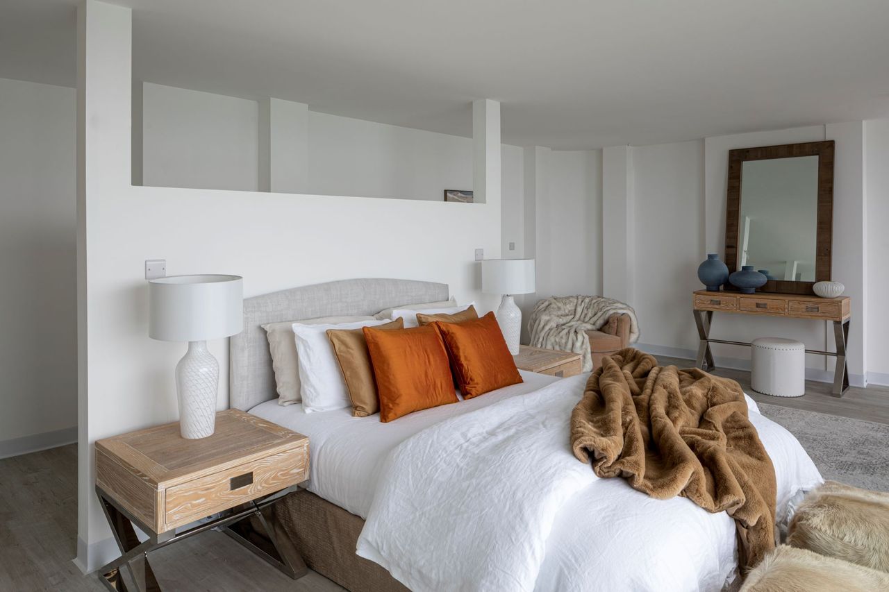 The bedroom has a large king-sized bed with white linen sheets and orange pillows and a room divider behind the bed leading into an open wardrobe. The room overlooks the sea