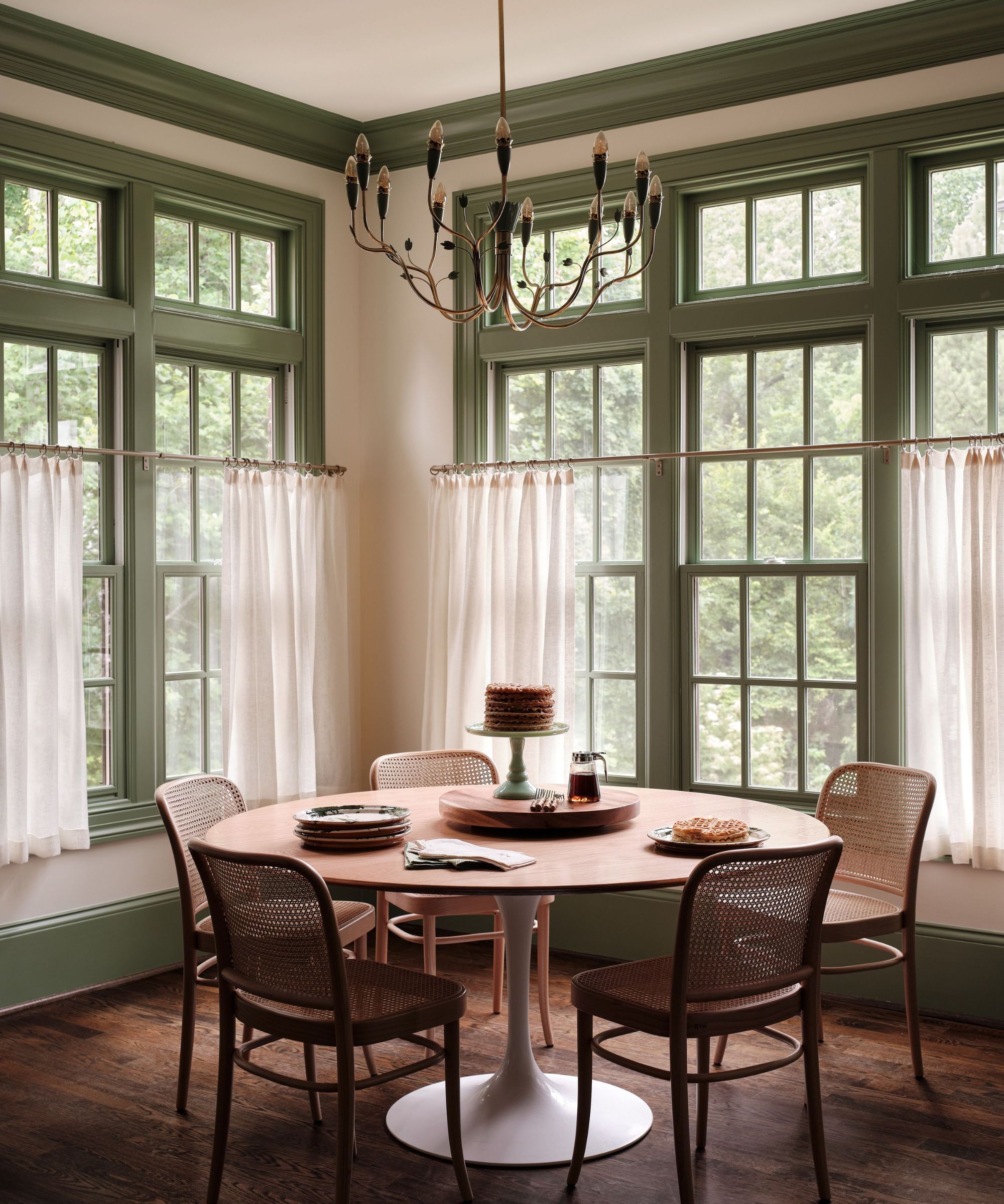A breakfast nook in a kitchen with large windows with green woodwork and cafe curtains, wood floors, a round dining table, and wooden chairs.
