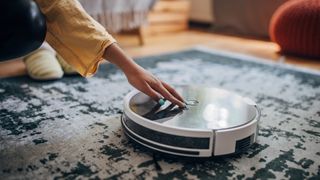 Close-up of a human hand pointing to a sleek robotic vacuum cleaner on a patterned rug in home interior