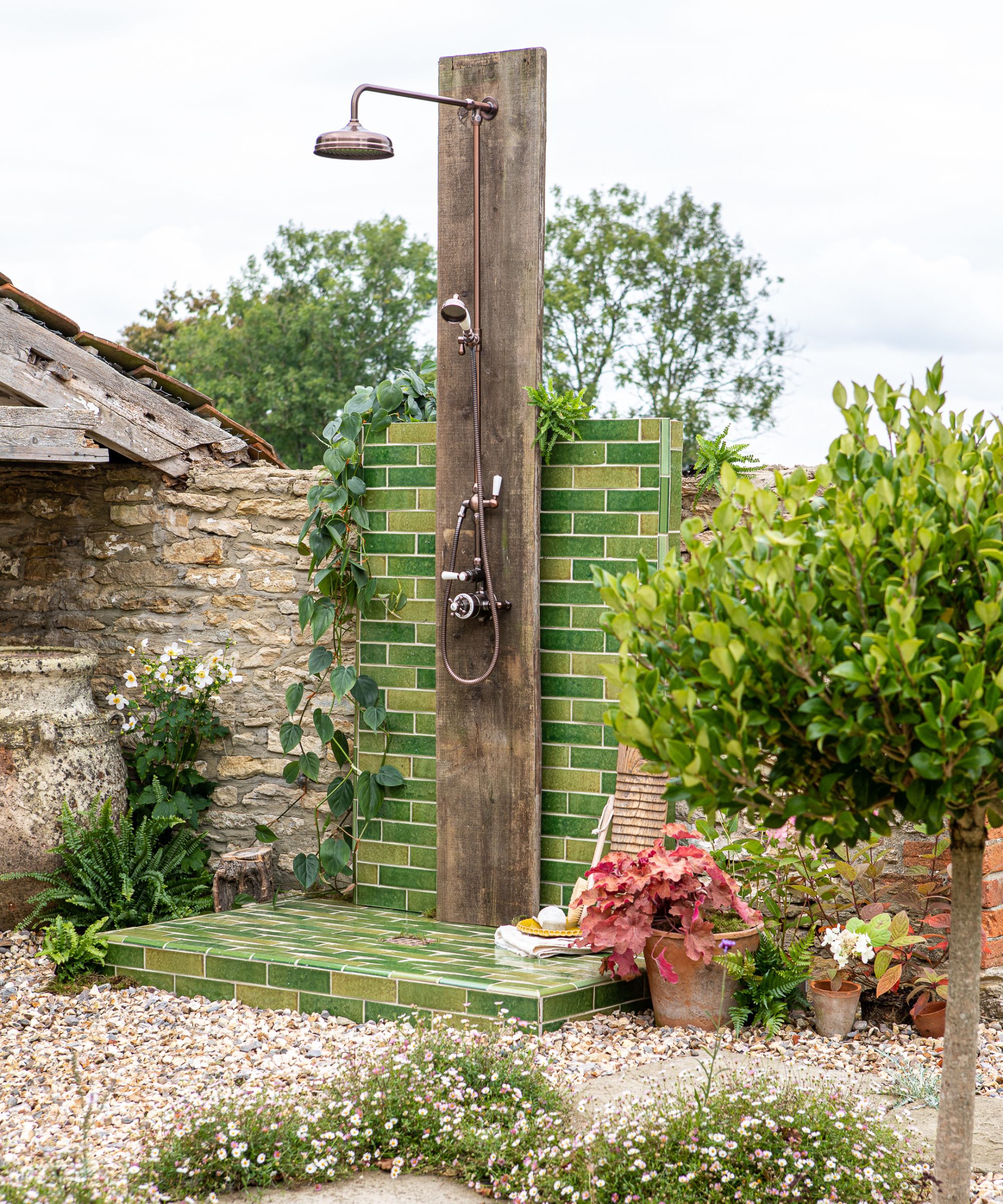A freestanding outdoor shower on a green zellige tiled platform, with a weathered wood upright post, antique copper rose head and controls, set among cottage garden planting and gravel.