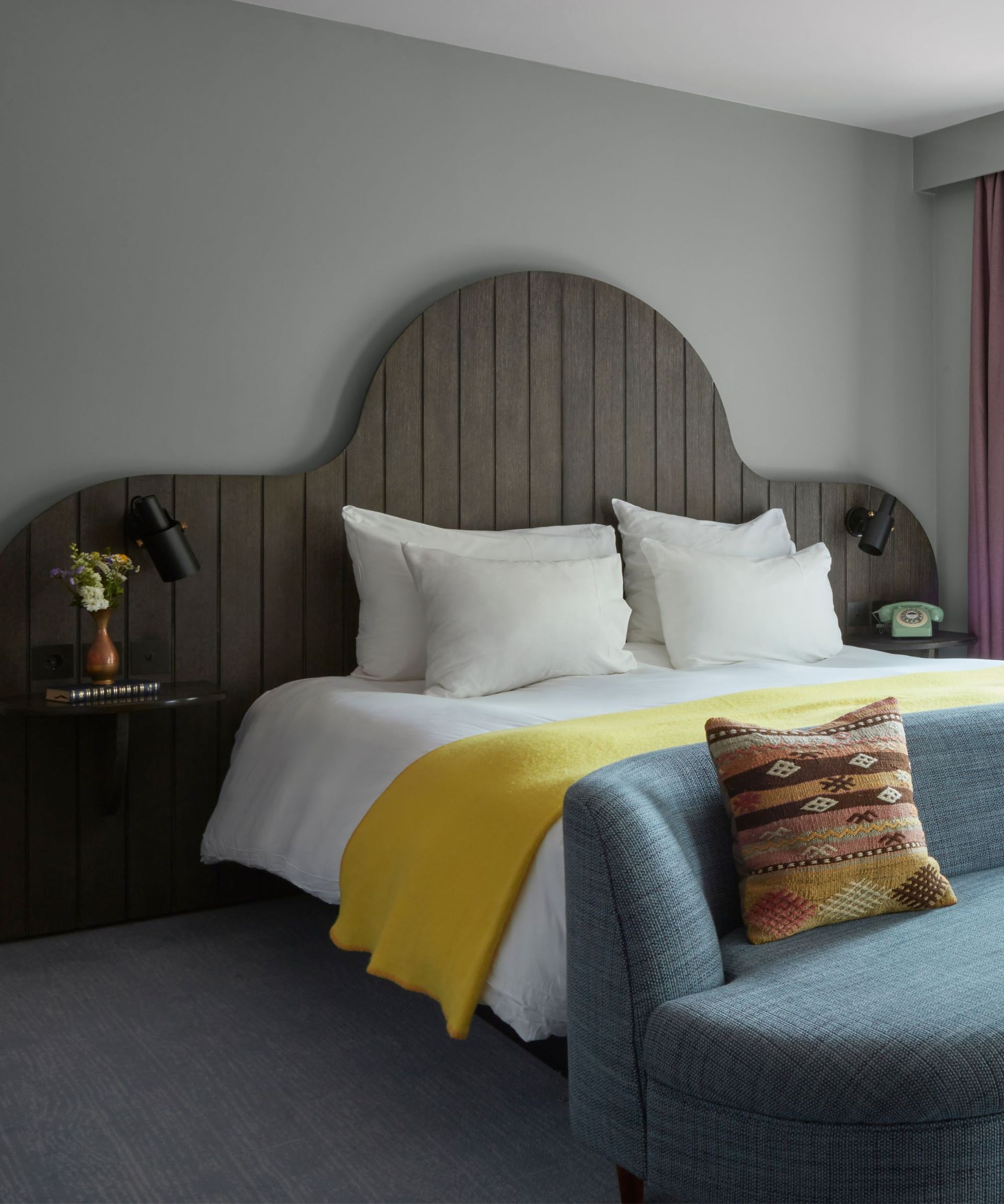 minimal hotel bedroom with gray walls and carpet and a large curved headboard with a light blue couch at the foot of the bed