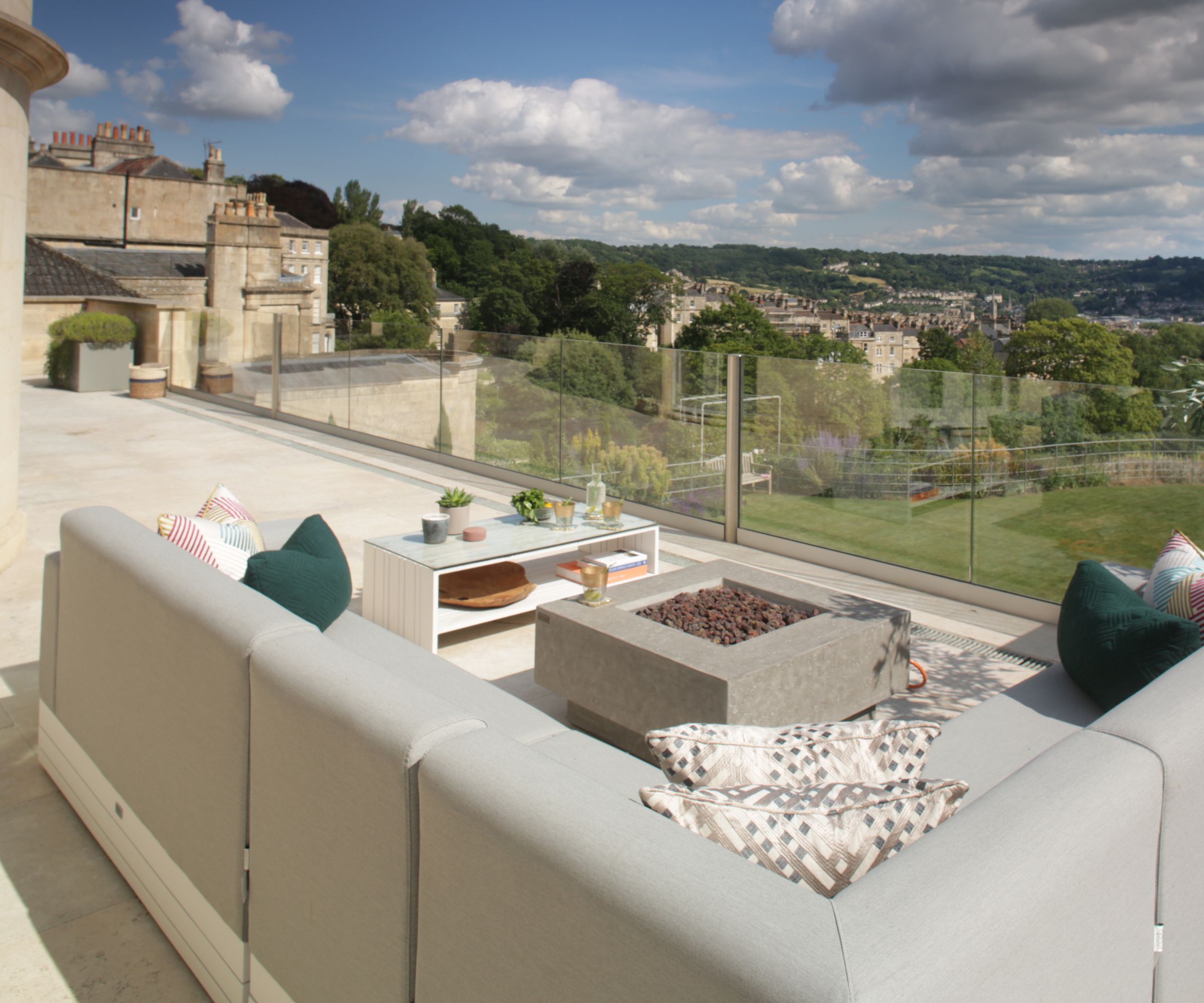 Grey outdoor sofas around square fire pit and coffee table, glass balustrade overlooking rooftops and hills.