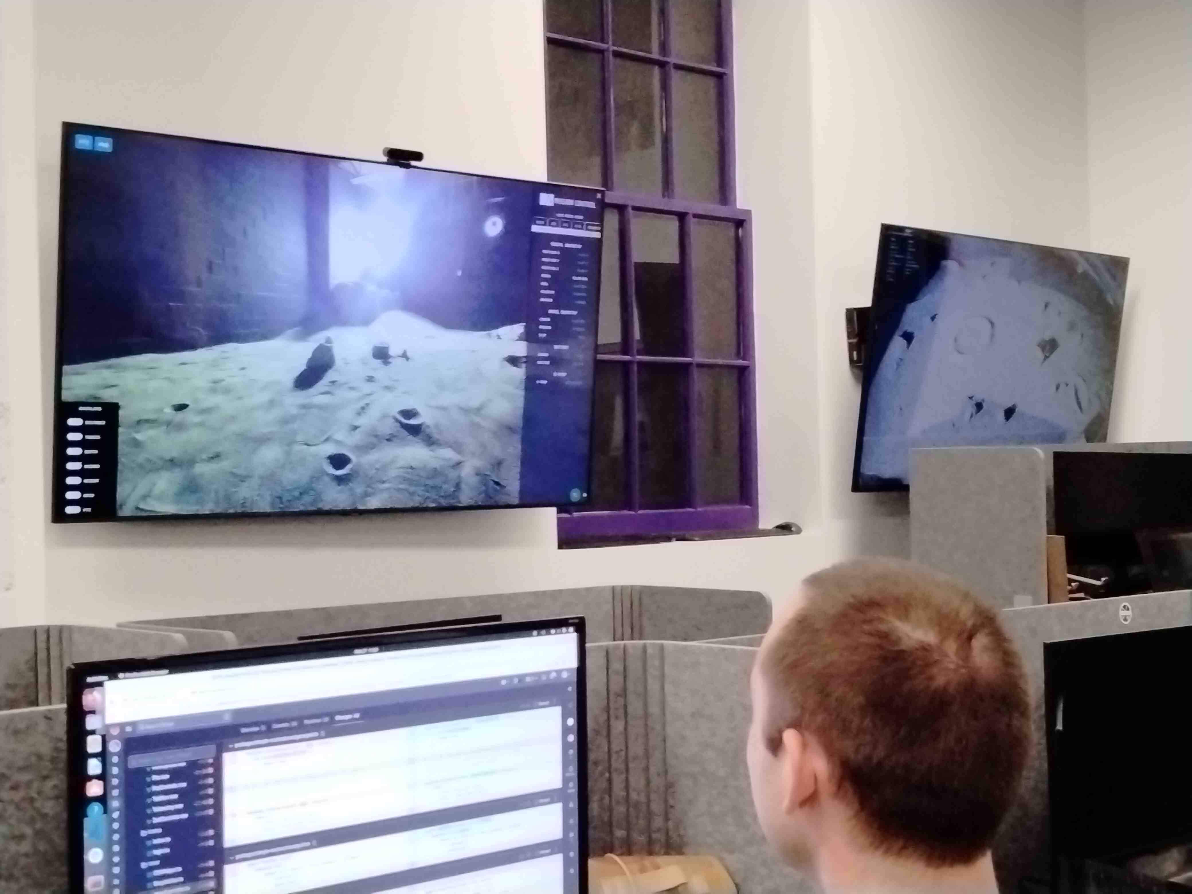 two television screens on the wall above a person working at a cubicle with a computer