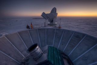 The sun sets behind BICEP2 telescope in the foreground. The South Pole Telescope stands in the background.