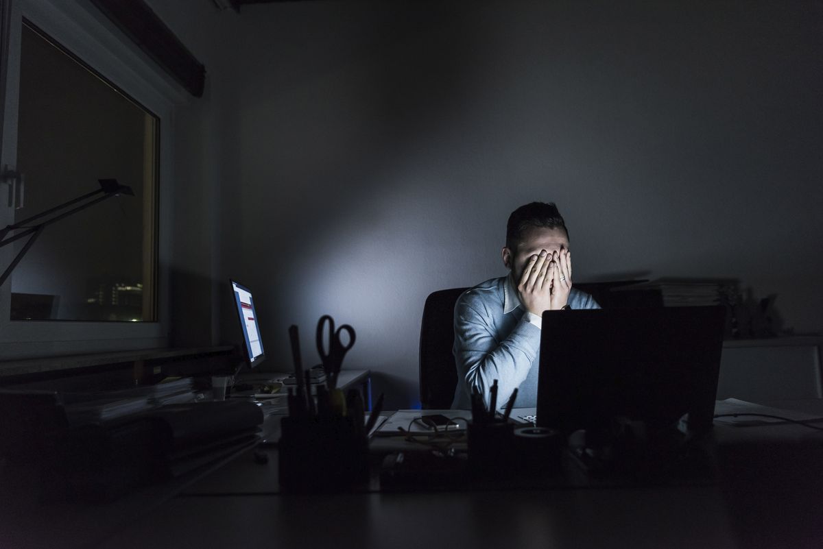 Exhausted worker sits behind desk at night in a darkened room in a case of work-related stress