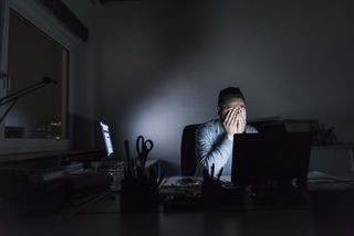 Stressed and exhausted office worker sits behind desk at night in a darkened room