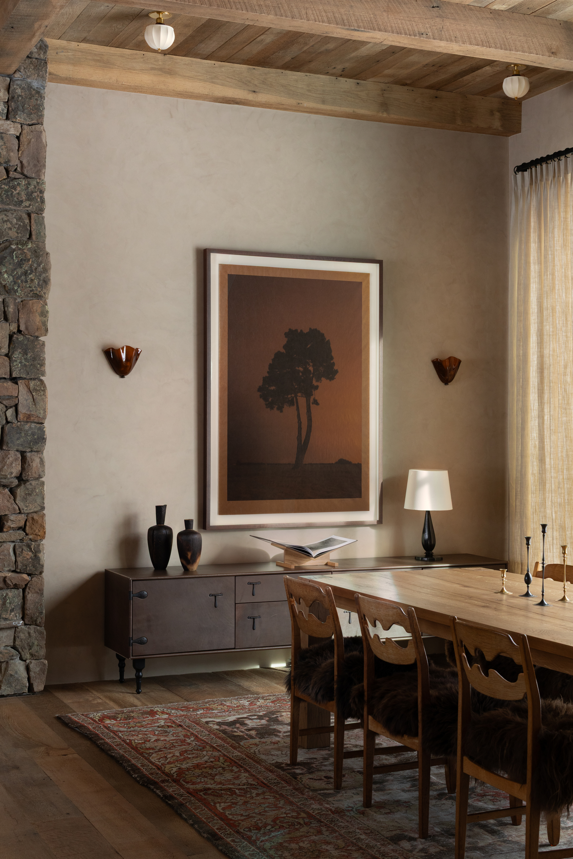 mountain house dining room with stacked stone wall, plastered walls, rug under timber dining table with vintage chairs, and a console along the wall styled with vases, a large artwork, wall sconces