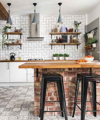 A brick kitchen island with a wooden counter and black metal stools behind it, with gray mosaic tiles on the floor, white subway tiles and wooden shelves on the wall, and three gray pendant lights on the ceiling