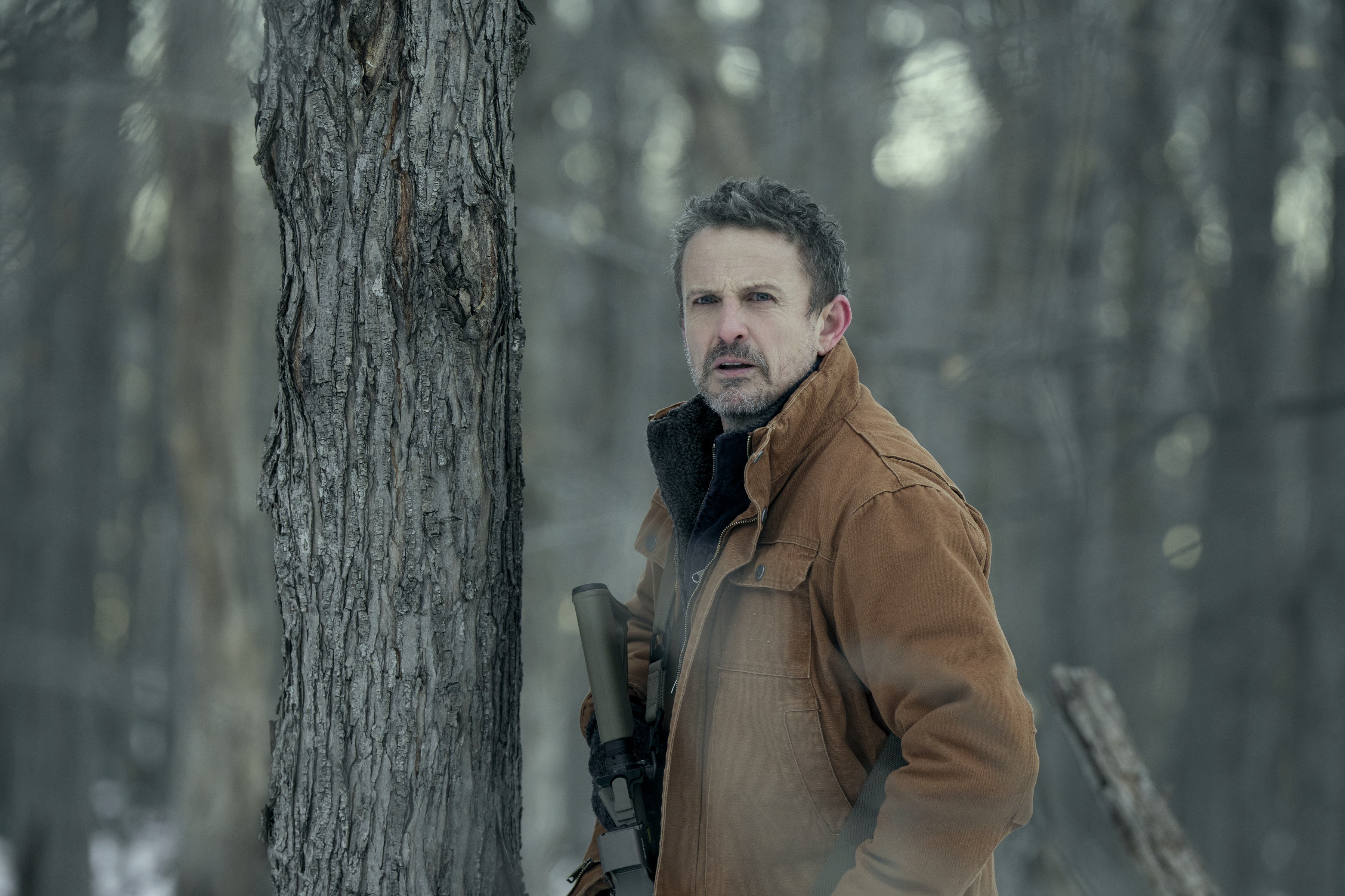 david lyons as adam wearing a brown jacket holding a gun and standing by a tree in a wintery woods in a still from the night agent season 3