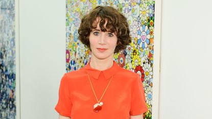 Miranda July wearing bright orange top and black pants in front of colorful canvas